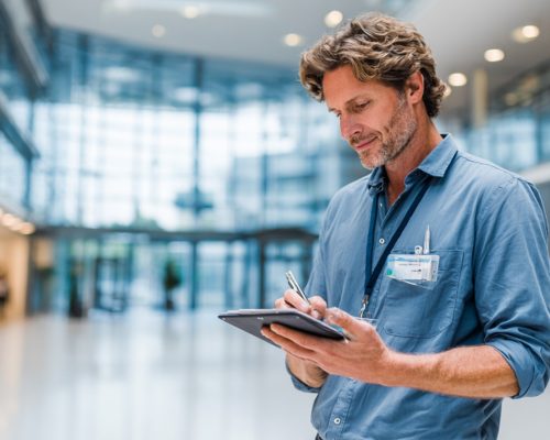I need a featured image for a blog post on how AI helps facility management technicians be more efficient. Ideally it would be a maintenance employee working with a device within a large institutional building (such as a hospital or a university campus). It should feel like the USA. Avoid commercial buildings (warehouses, retail stores) ideally it will be a large room like an atrium with a lot of natural light and foot traffic. Used device, not a clipboard. --ar 3:2 --v 7 Job ID: d3624a0a-4421-4e41-abb5-d382fab18222